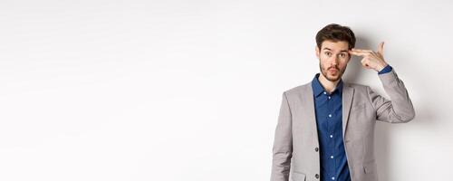 Annoyed guy in business suit shoot himself with hand gun near head, look distressed and tired after work, standing on white background photo