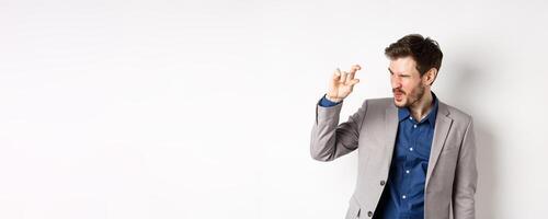 Businessman trying to see something tiny, showing little thing size with fingers, standing in suit on white background photo