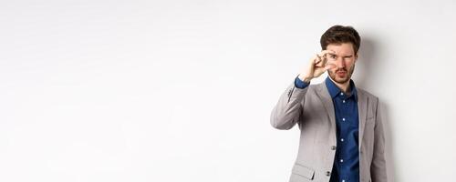 Businessman trying to see something tiny, showing small thing size with fingers, standing in suit on white background photo