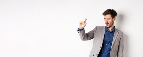 Businessman trying to see something small, showing little thing size with fingers, standing in suit on white background photo