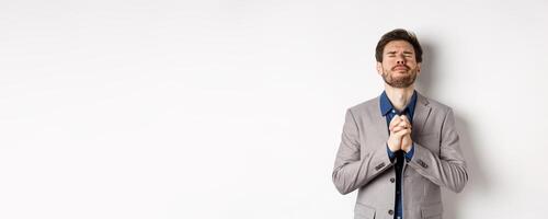 Nervous male entrepreneur in suit begging god, holding hands in pray with pleading expression, close eyes and make wish, asking for help, standing on white background photo