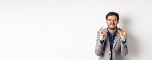 Angry and tensed businessman wants to kill someone, clenching hands and teeth, looking mad at camera, going to strangle person, standing on white background photo