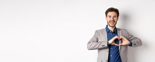 Passionate and romantic man in suit falling in love, showing heart sign and smiling at lover, standing on white background photo