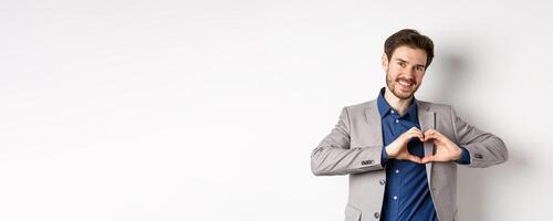 Romantic man in suit showing heart sign and smiling, love his girlfriend, standing on white background, making confession to lover photo