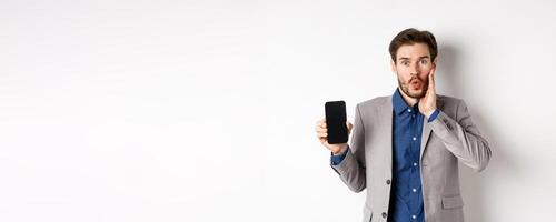 E-commerce and online shopping concept. Excited man gasping amaze and showing empty smartphone screen, wearing business suit, white background photo