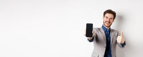E-commerce and online shopping concept. Cheerful businessman in suit showing thumbs up and empty smartphone screen, recommending app, standing on white background photo
