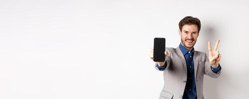 E-commerce and online shopping concept. Cheerful man in business suit showing v-sign and empty mobile phone screen, demonstrate smartphone app, white background photo