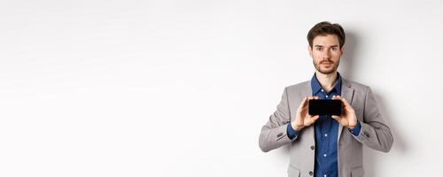 E-commerce and online shopping concept. Serious young man in suit showing empty smartphone screen horizontal, standing on white background photo