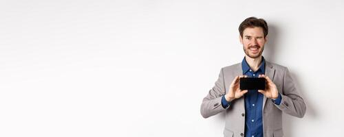 E-commerce and online shopping concept. Cheeky male model in suit winking and showing empty smartphone screen, smiling happy at camera, white background photo