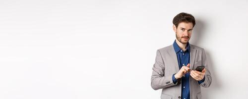 Handsome bearded male model in suit using mobile phone, smiling pleased at camera, white background photo