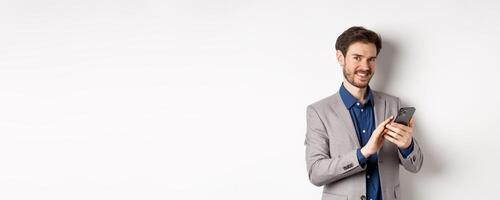 Smiling successful business man using smartphone on white background, standing in suit photo