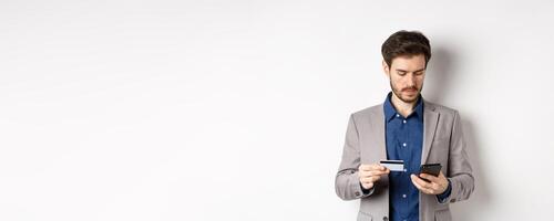 Handsome businessman paying online with credit card and smartphone, standing on white background in suit photo