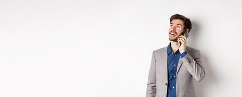 Carefree young man talking on mobile phone and smiling, looking up with dreamy face, standing in suit on white background photo