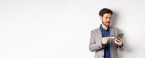Online shopping. Smiling businessman paying with credit card on smartphone, sending money, standing in suit on white background photo