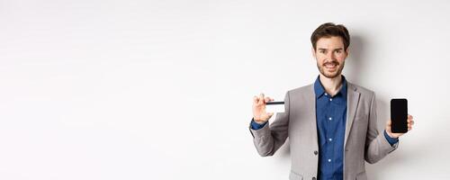 Online shopping. Smiling business man in suit showing plastic credit card with empty smartphone screen, standing against white background photo