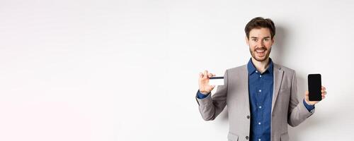 Online shopping. Smiling business man in suit showing plastic credit card with empty smartphone screen, standing against white background photo