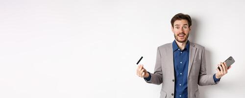 Online shopping. Surprised businessman holding plastic credit card and smartphone, making money in internet, standing in suit against white background photo