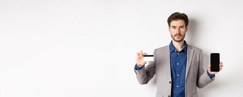 Online shopping. Serious bearded man in business suit showing plastic credit card with empty smartphone screen, standing against white background photo