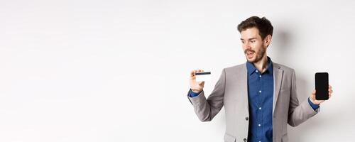 Online shopping. Smiling business man in suit showing plastic credit card with empty smartphone screen, standing against white background photo