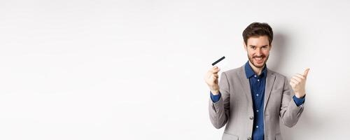 Happy bank client in business suit showing plastic credit card and thumb up, smiling satisfied, standing on white background photo