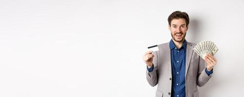 Happy successful businessman in suit showing money and credit card, smiling at camera, white background photo