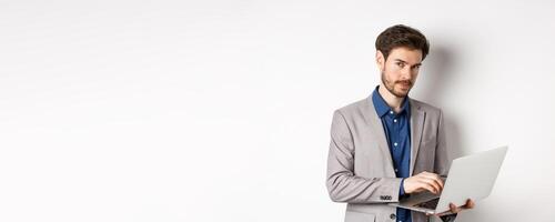 Handsome businessman in suit working on laptop, looking at camera confident, standing against white background photo