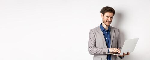 Successful smiling businessman working on laptop and looking happy at camera, standing in grey suit on white background photo