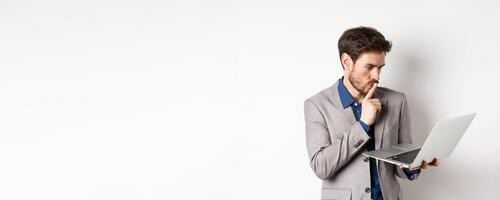 Pensive businessman working on laptop and thinking, looking thoughtful at computer screen, standing in suit on white background photo