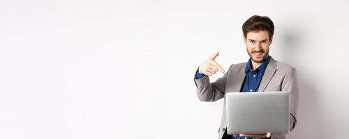 Confident young male entrepreneur working online with laptop, pointing at computer and smiling, standing on white background photo