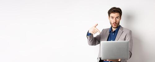 Excited handsome man asking to look here, pointing at laptop screen with amazed face, standing in business suit against white background photo