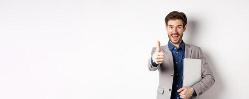 Good job. Happy smiling business man in suit holding laptop, showing thumb up to praise you, nice work gesture, standing on white background photo