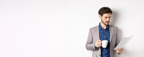 Boss reading documents and drinking morning coffee from mug, looking at paper serious, standing in suit on white background photo