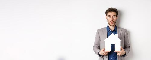 Real estate. Amazed caucasian man in suit checking out special offer for property, standing with paper house cutout and say wow at camera, white background photo