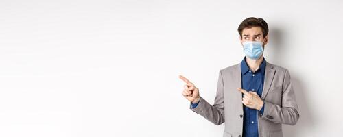Covid-19, pandemic and business concept. Hesitant businessman in medical mask and suit looking, pointing left with doubtful face, standing on white background photo