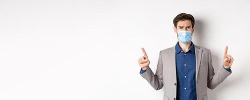Covid-19, pandemic and business concept. Disappointed frowning guy in suit and medical mask pointing fingers up, complaining on unfair situation, white background photo