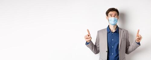 Covid-19, pandemic and business concept. Handsome businessman in medical mask and suit, looking and pointing up with dreamy face, white background photo