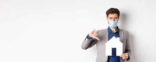 Real estate and covid-19 concept. Disappointed guy in medical mask and suit showing paper house cutout with thumbs-down, complaining on agency, white background photo