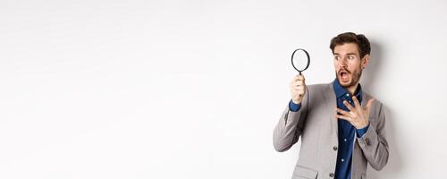 Shocked guy look through mafnifying glass with startled face, gasping and holding hand on chest scared, standing on white background photo