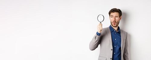 Excited funny guy in suit look through magnifying glass aside, checking out awesome thing, found something, white background photo
