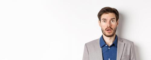 Close up portrait of shocked caucasian man in suit drop jaw, looking speechless at camera, white background photo
