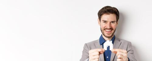 Real estate and insurance concept. Handsome man in suit smiling, showing small paper house cutout, standing on white background photo