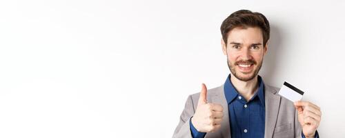 Shopping. Handsome satisfied client showing thumbs up and plastic credit card, smiling pleased, white background photo