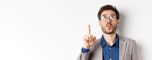 Excited handsome man in glasses and business suit, look up and pointing at top advertisement, exclaim wow with amazed expression, white background photo