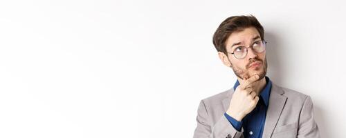 Thoughtful businessman with beard and glasses making decision, looking up and thinking, standing in suit with pensive face, white background photo