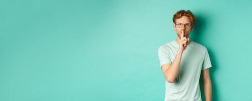 Serious redhead man hushing, tell to be quiet, making shush gesture and looking at camera, standing in t-shirt over turquoise background photo