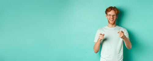Handsome young man with ginger hair, wearing glasses and t-shirt, pointing finger at camera and smiling, choosing you, congratulating or praising, standing over mint background photo