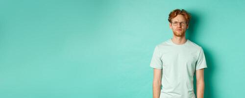 Confused young redhead man in glasses staring at camera puzzled, squinting perplexed, cant understand something, standing over mint background photo