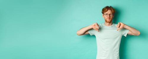 Disappointed caucasian guy with red messy hair and glasses showing thumbs down, dislike something, frowning upset and displeased, standing over mint background photo