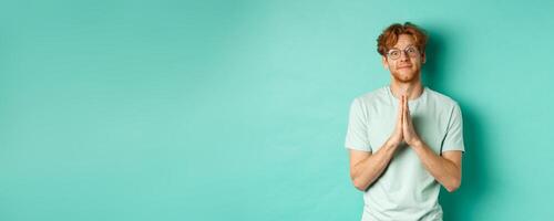 Cute redhead man asking for help, saying please with namaste gesture, pleading or begging for favour, looking silly at camera, standing over mint background photo