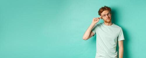 Skeptical redhead man scolding someone stupid or crazy, pointing finger at head and staring at camera, standing over mint background photo